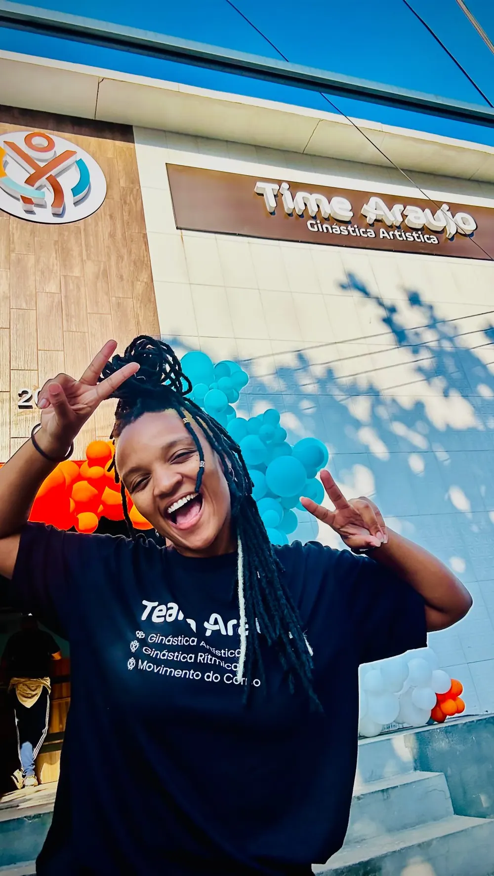 Professora sorri em frente à fachada do Time Araújo, decorada com balões laranja e azuis
